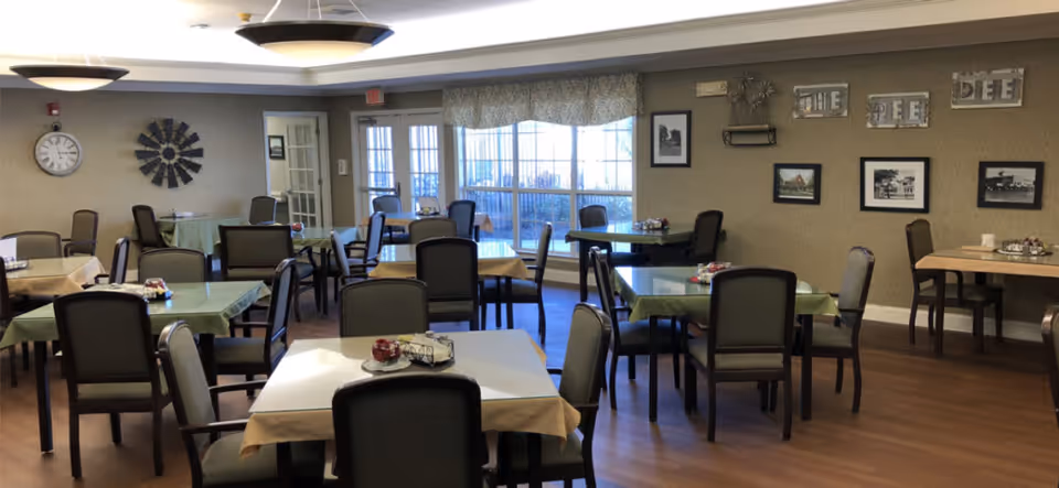 A dining room with multiple tables covered with green and beige tablecloths, each surrounded by chairs. The room has large windows with curtains letting in natural light. The walls are decorated with framed pictures and a clock. The floor is wooden, and ceiling lights provide additional illumination.