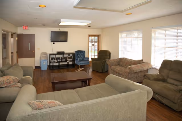 Bright communal living room with sofas and armchairs arranged around a coffee table, a wall-mounted TV, and windows with blinds.