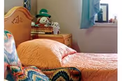 A cozy bedroom with a bed covered in an orange quilt. Next to the bed is a wooden nightstand with a basket and two stuffed animals on top. A window with a light blue curtain is visible in the background, and part of a patterned armchair is seen in the foreground.