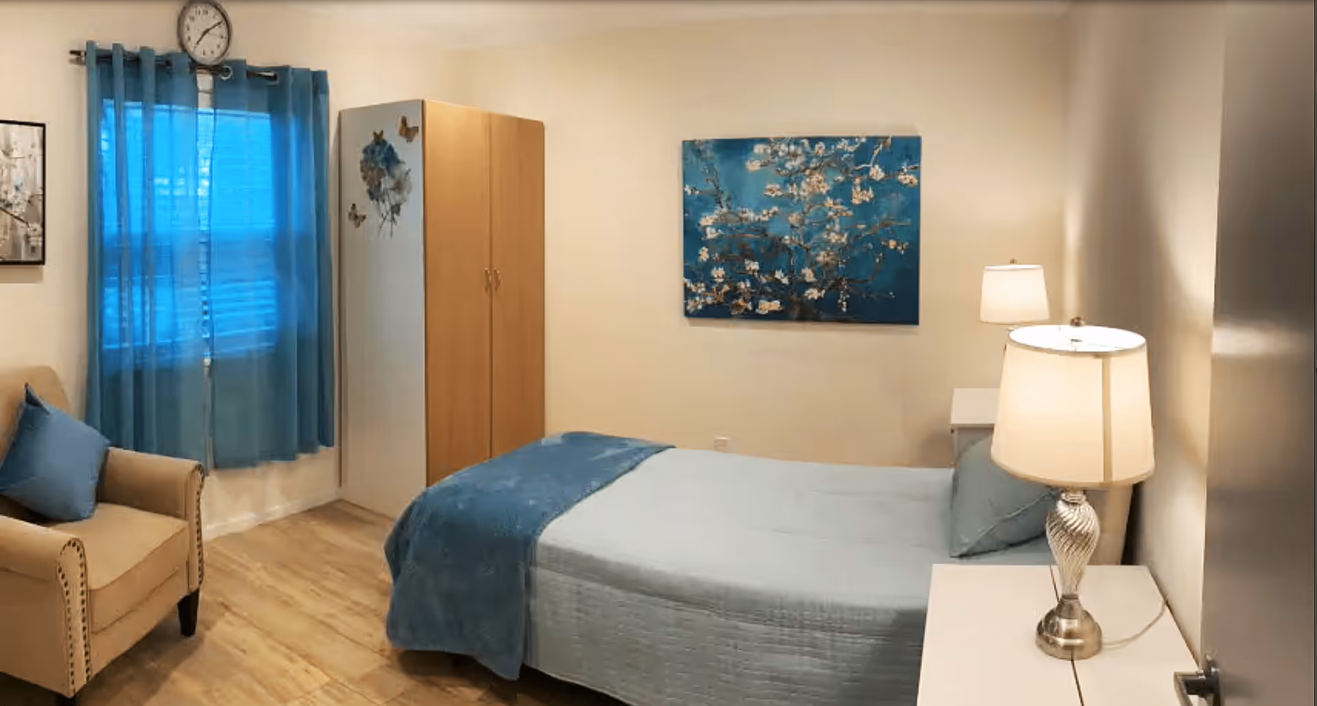 Well-lit bedroom with a made bed, bedside tables and lamps, an armchair, wardrobe, blue curtains and wall art.