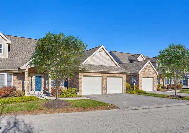 Attached townhome-style units with stone facades, multiple garage doors, front entrances, small trees and driveways under a clear blue sky.