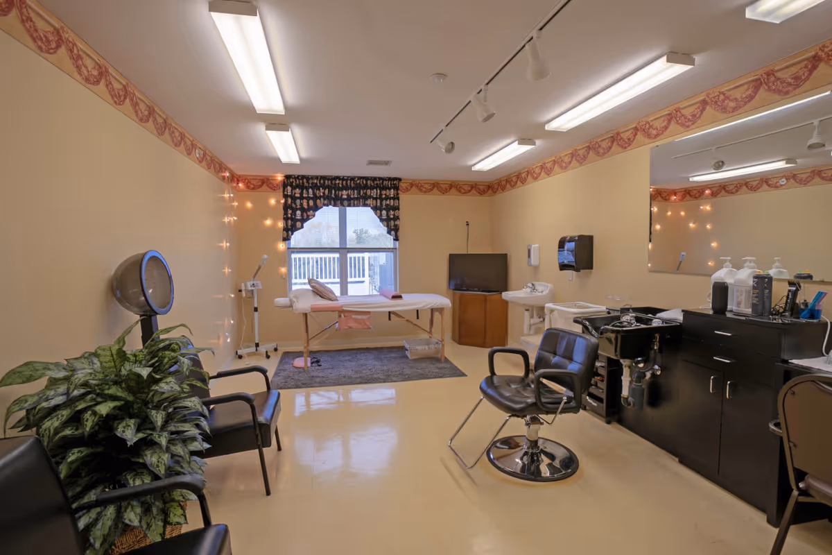 A well-lit beauty salon room with beige walls and a decorative border near the ceiling. The room features a black salon chair in front of a black sink and counter with various hair care products. There is a hair dryer chair, a couple of black waiting chairs, a large mirror on the wall, and a massage or treatment table near a window with a dark patterned valance. The floor is light-colored and shiny, and there are string lights hanging on one wall.