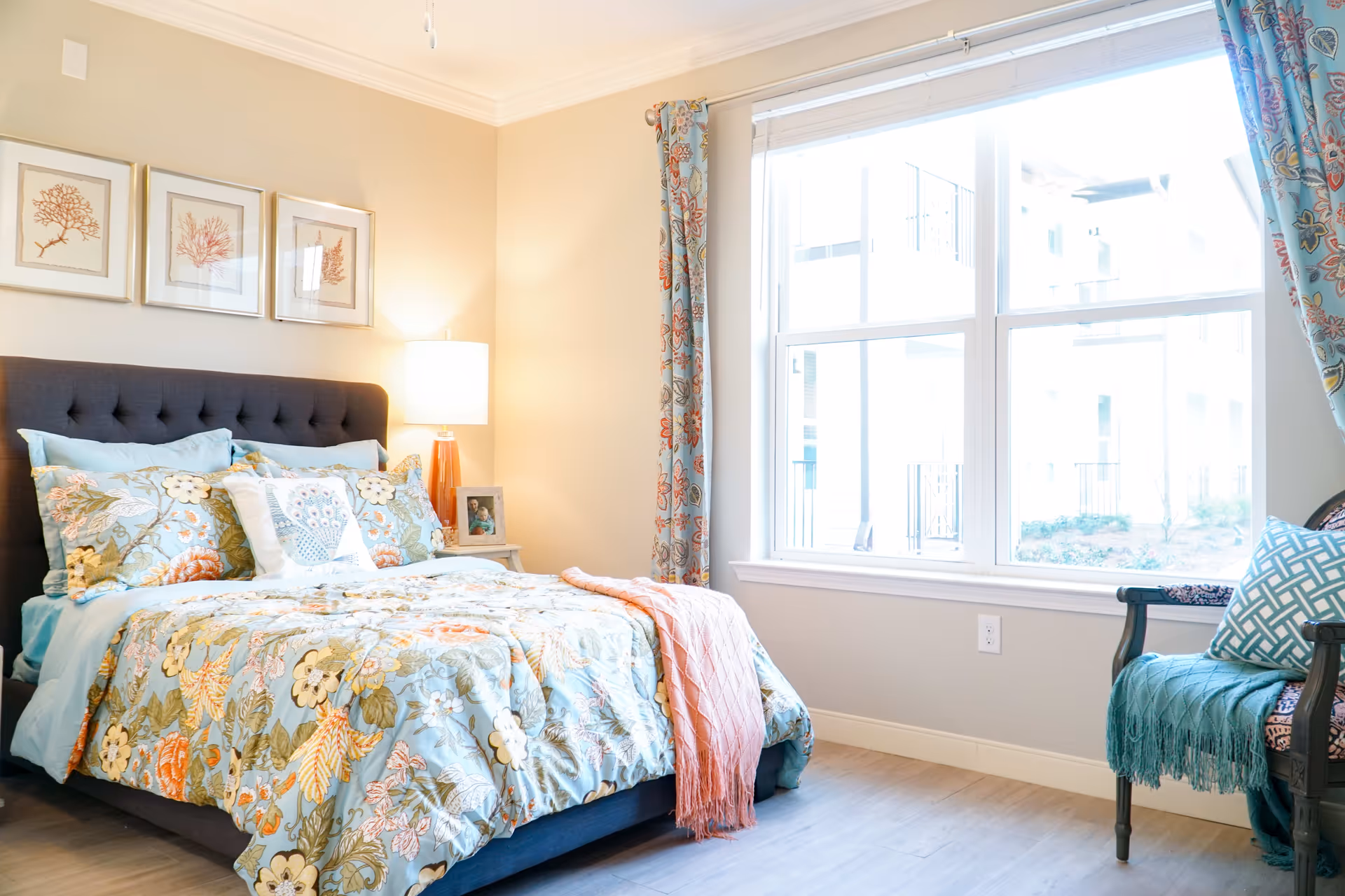 A bright and cozy bedroom with a large window letting in natural light. The bed has a dark upholstered headboard and is dressed with floral patterned bedding in shades of blue, green, and orange. There is a bedside table with a lamp and a framed photo. A chair with a blue throw and patterned pillow is placed near the window. Three framed coral-themed artworks hang on the wall above the bed.