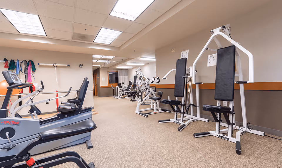 Interior view of a fitness room with various exercise machines including stationary bikes and weight machines. The room has beige walls, carpeted floor, and fluorescent ceiling lights. Exercise bands and a large orange exercise ball are visible on the left wall.
