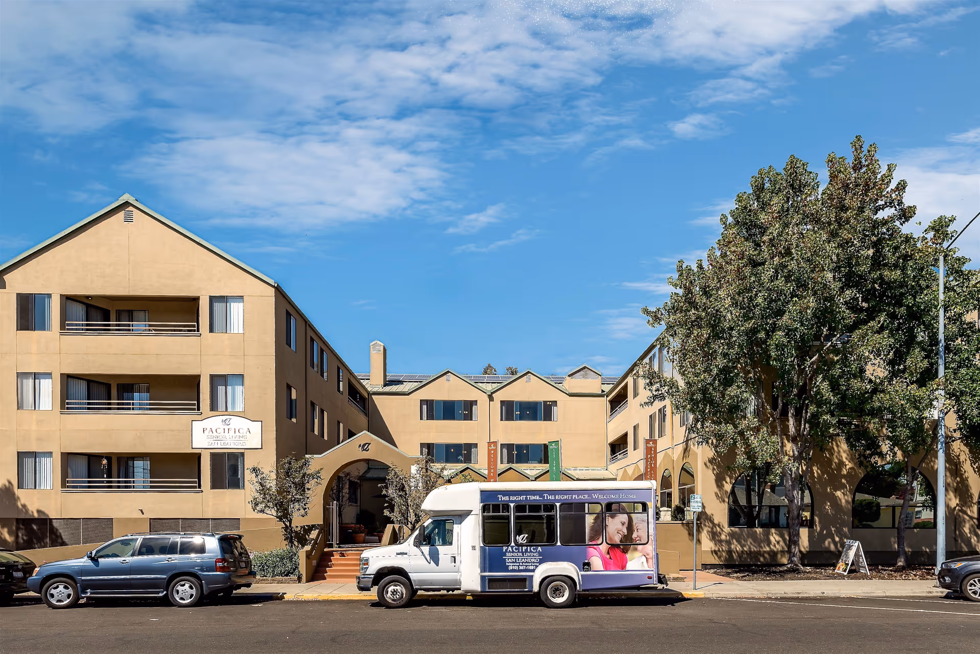 Exterior view of a multi-story senior living facility building with beige walls and multiple windows. A white shuttle bus with Pacifica Senior Living branding is parked in front of the building. There are several trees and parked cars along the street under a partly cloudy blue sky.