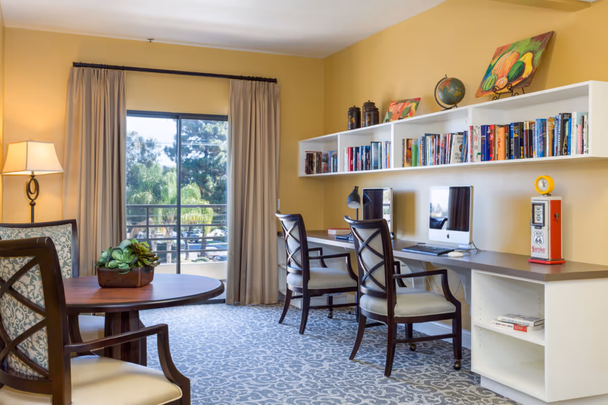 A cozy interior room with a round wooden table and two upholstered chairs on the left. On the right, there is a long desk with two chairs, two desktop computers, a desk lamp, and shelves above filled with books and decorative items including a globe and colorful paintings. A large window with beige curtains lets in natural light and shows trees outside.
