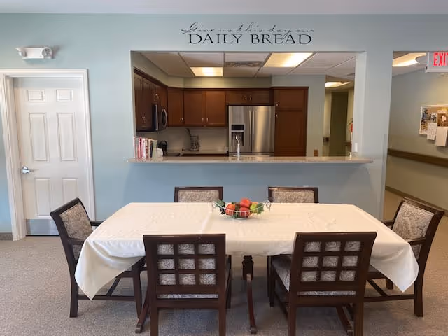 Dining area with a rectangular table covered with a white tablecloth and six wooden chairs with cushioned seats. A bowl of fruit is placed in the center of the table. Behind the table is a kitchen with wooden cabinets, a stainless steel refrigerator, and a countertop. Above the kitchen opening, there is a wall decal that reads 'Give us this day our DAILY BREAD'.