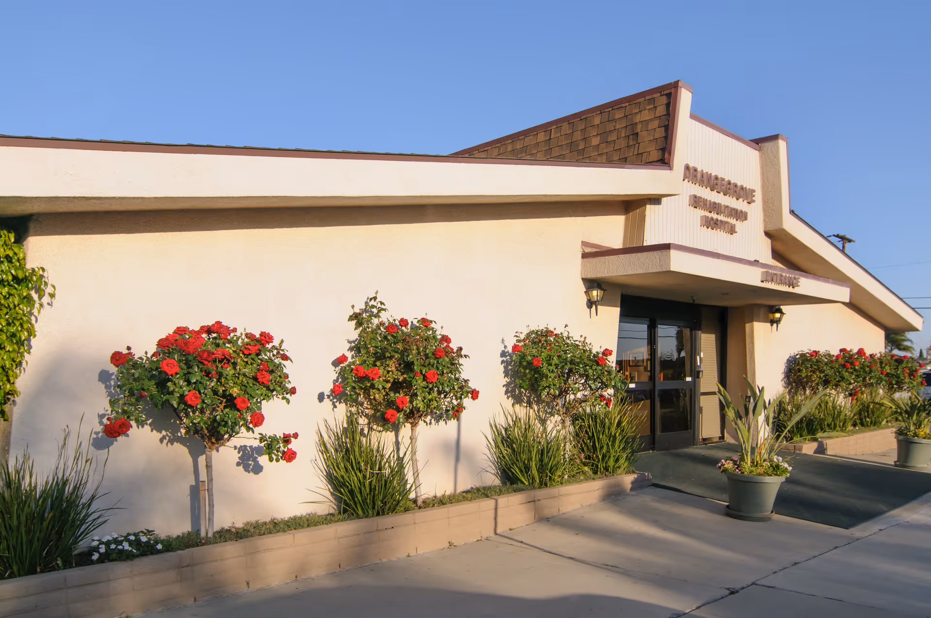 Exterior view of Orangegrove Rehabilitation Hospital building with a beige facade, a sloped roof with brown shingles, and an entrance with double glass doors. There are red rose bushes and other plants along the front of the building under a clear blue sky.