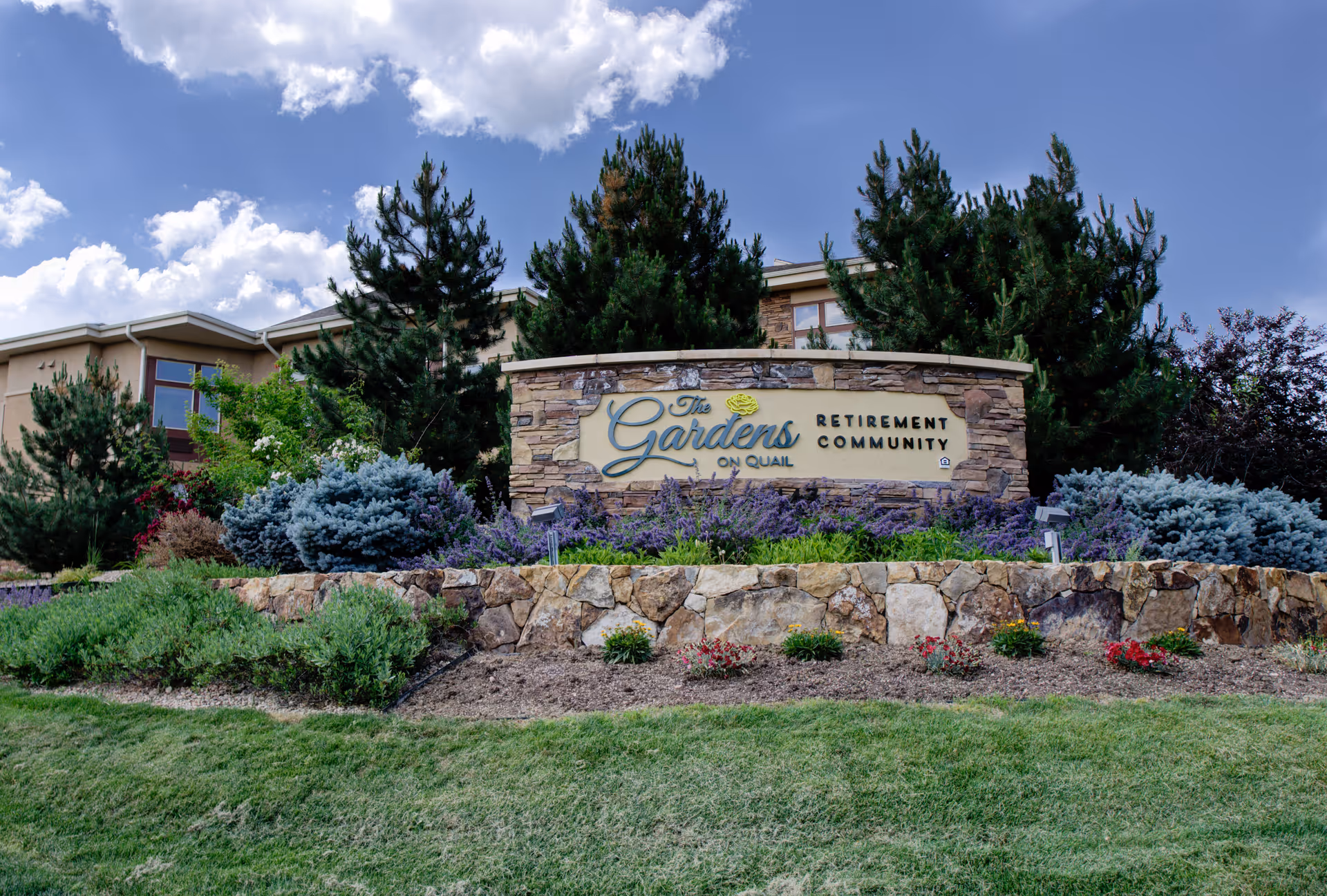 Stone sign for The Gardens on Quail Retirement Community surrounded by landscaped bushes, flowers, and trees with a building partially visible in the background under a partly cloudy sky.
