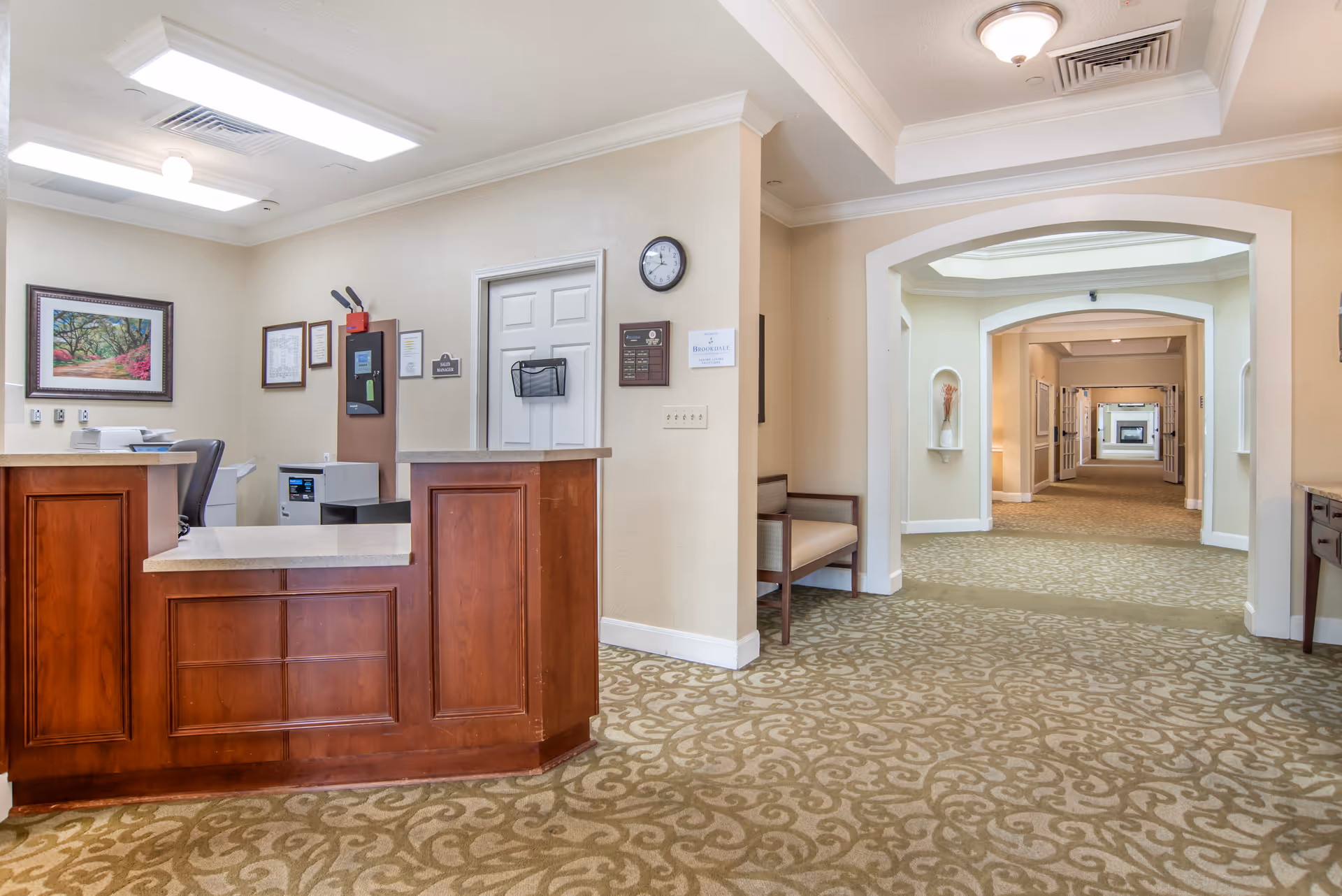 Reception area with a wooden front desk, office chair, and printer. The walls are beige with framed pictures and a clock. A hallway with patterned carpet and arched doorways extends into the distance, with a bench seating area on the right side.