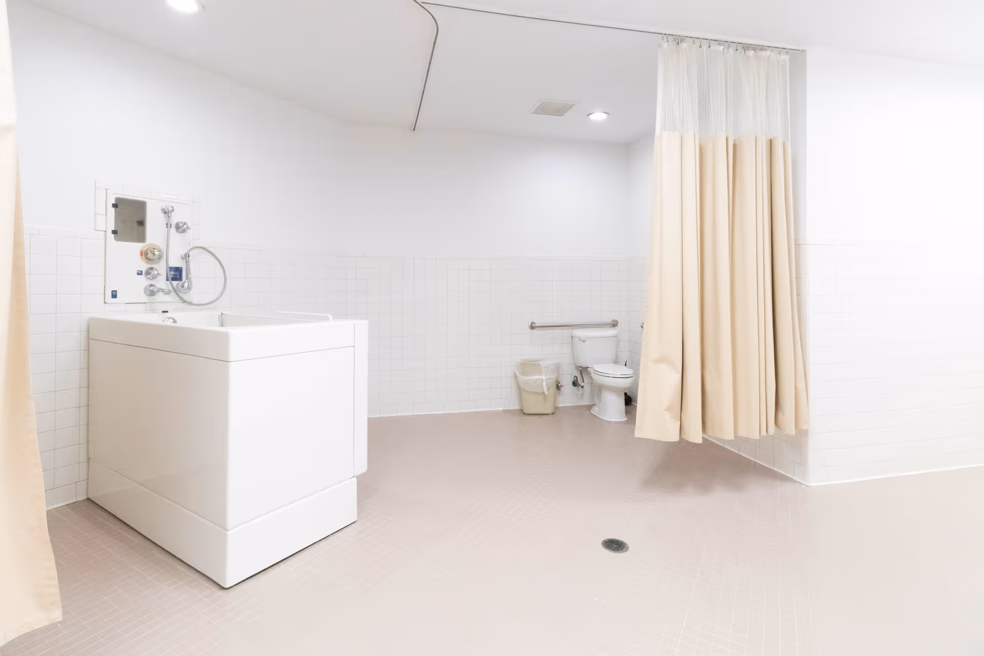 A spacious, clean bathroom with a large white walk-in bathtub on the left, a toilet with a grab bar on the wall behind it, a trash bin, and a beige privacy curtain hanging from the ceiling. The walls are tiled halfway up with white tiles, and the floor is covered with light-colored tiles.