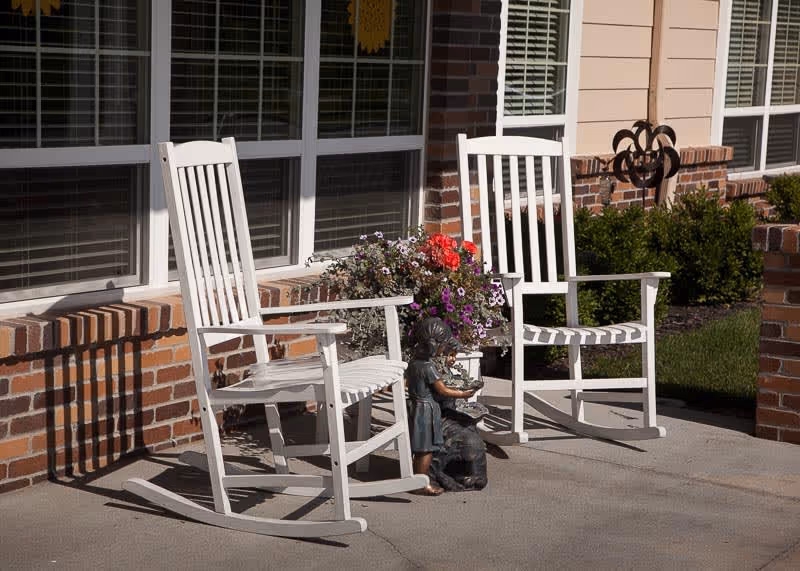 Two white wooden rocking chairs on a concrete porch outside a brick and siding building, with a flower pot containing colorful flowers placed between them and a small statue of a girl with a dog in front of the chairs.