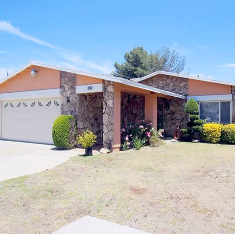 Single-story residential building with a stone facade and peach-colored walls, featuring a two-car garage on the left and a covered entrance with plants and shrubs in front. The sky is clear and blue.