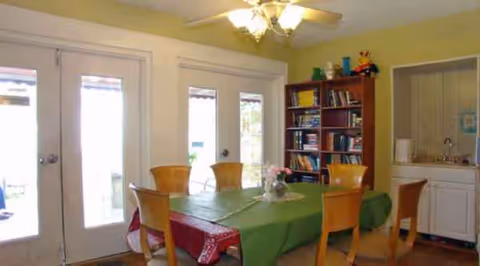 Senior living dining room featuring a green-clothed table, wooden chairs, bookshelf, and kitchenette.