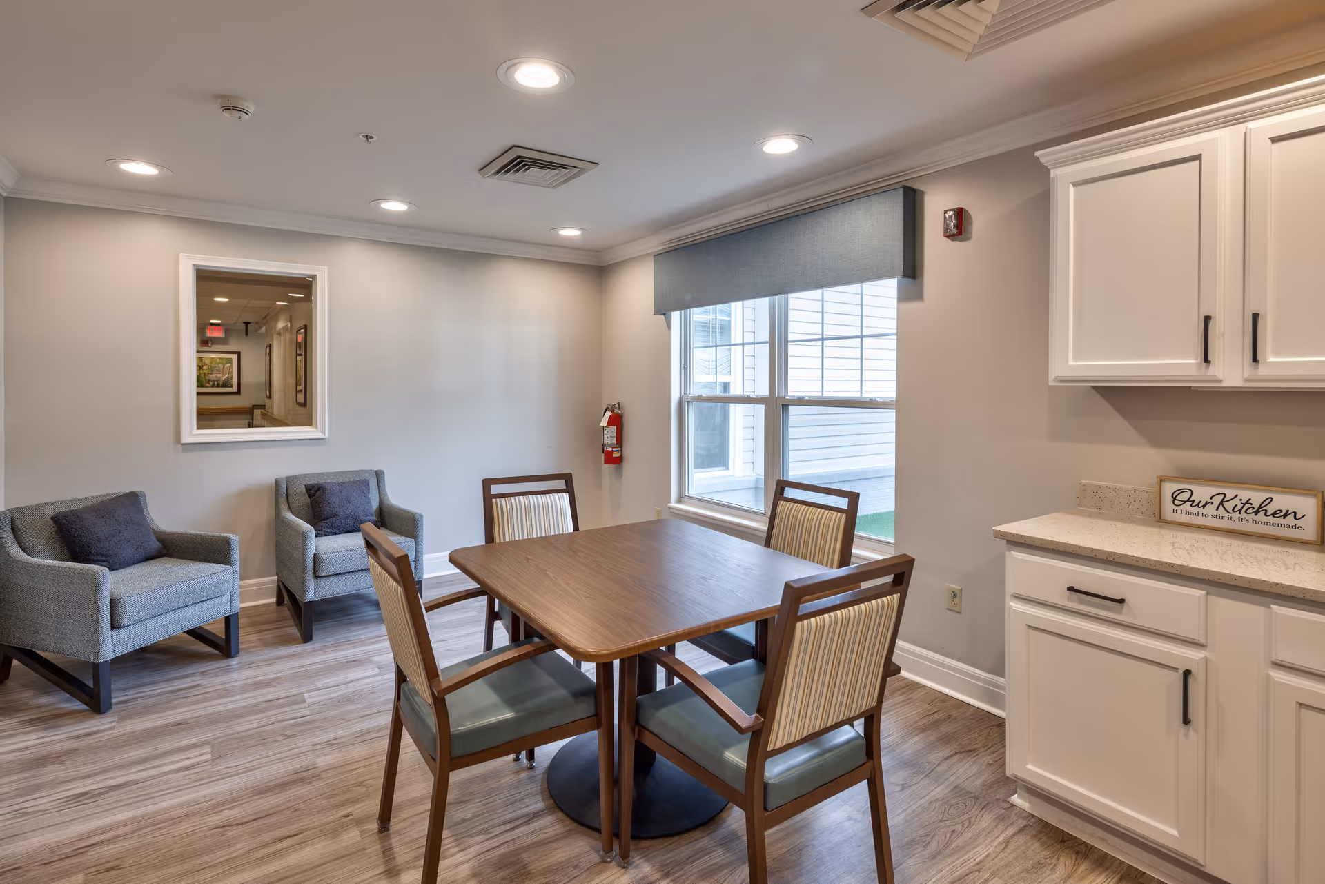 A cozy interior room with a wooden table surrounded by four chairs with striped upholstery. Two gray armchairs with dark cushions are placed against a light gray wall with a window covered by a blue valance. White cabinets and a countertop are visible on the right side, with a small sign that reads 'Our Kitchen if I had to stir it, it's homemade.' The room has recessed ceiling lights and wood-patterned flooring.