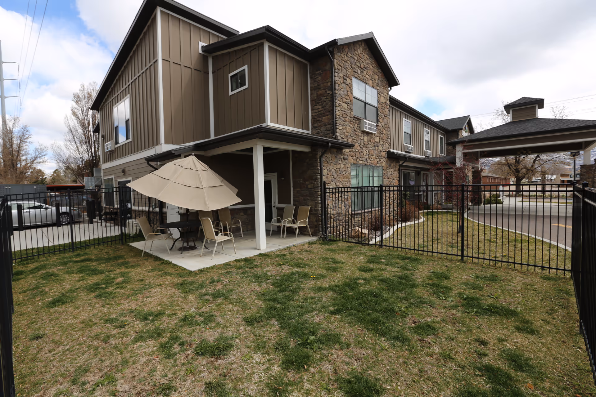 Two-story stone-and-siding memory care building with a fenced grassy yard, a covered patio with umbrella and chairs, and a drive-up canopy.