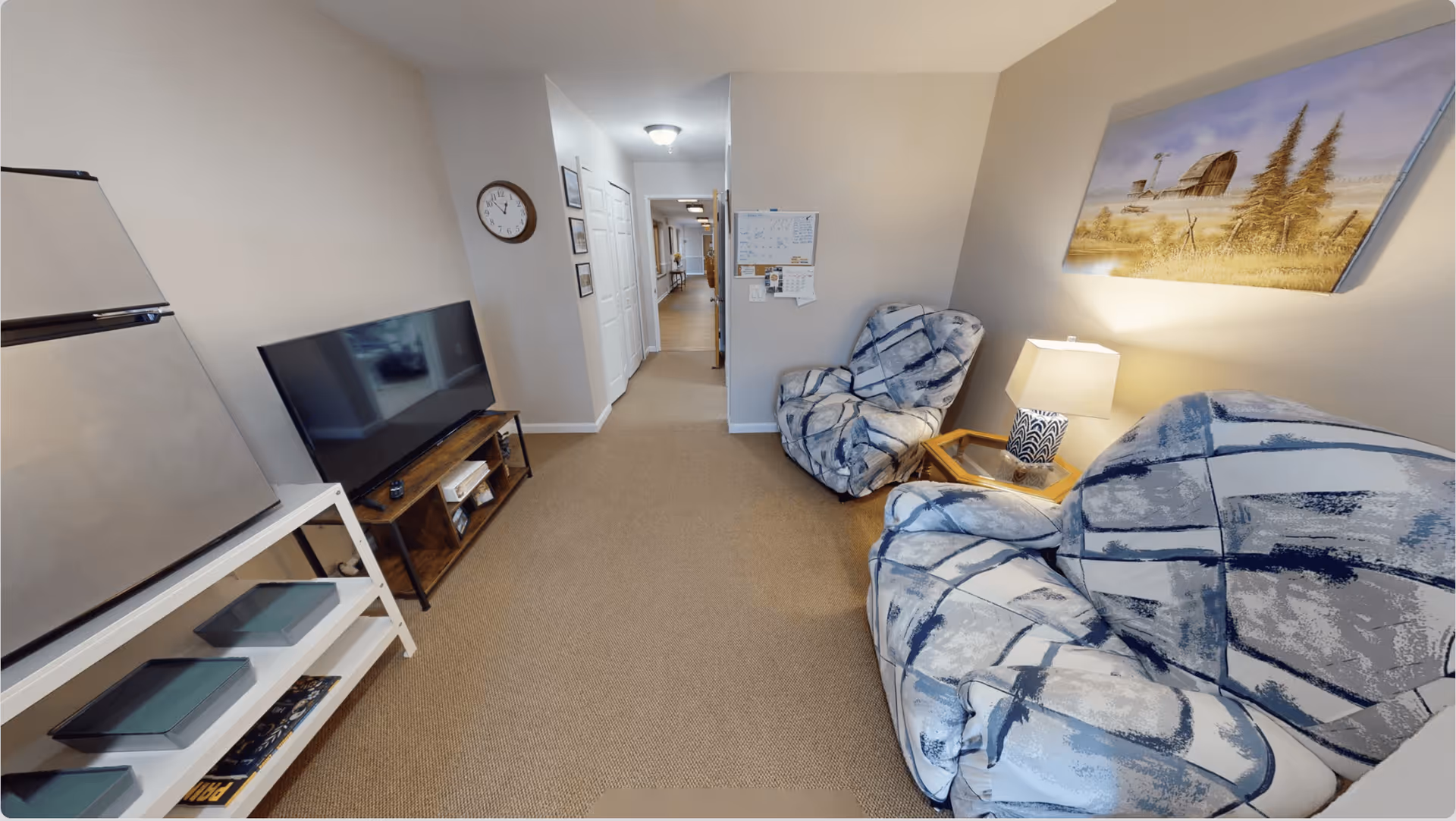 A cozy living room area in Aspen Assisted Living featuring two patterned armchairs, a small wooden side table with a lamp, a flat-screen TV on a wooden stand, a refrigerator, and a white shelving unit with trays. The walls are light-colored, decorated with a clock, framed pictures, a calendar, and a painting of a barn and trees. A hallway extends from the living room area.