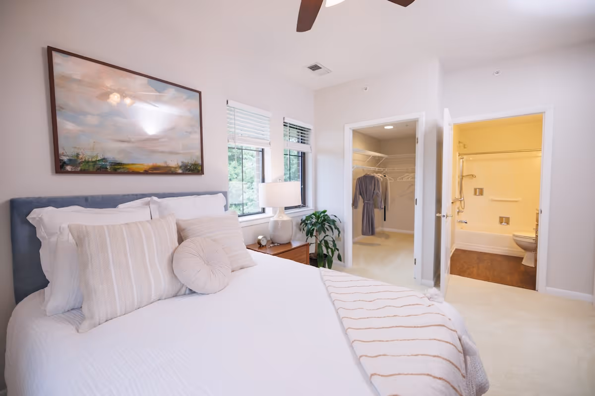 A bright bedroom with a neatly made bed featuring white and beige pillows and a striped throw blanket. There is a wooden nightstand with a lamp and a small plant next to two windows with white blinds. On the wall above the bed is a large framed painting of a landscape. Two open doors reveal a walk-in closet with hanging clothes and a bathroom with a bathtub, toilet, and grab bars.