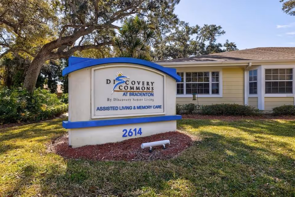 Outdoor view of a sign for Discovery Commons At Bradenton, an assisted living and memory care facility, with a yellow building and windows in the background surrounded by trees and grass.