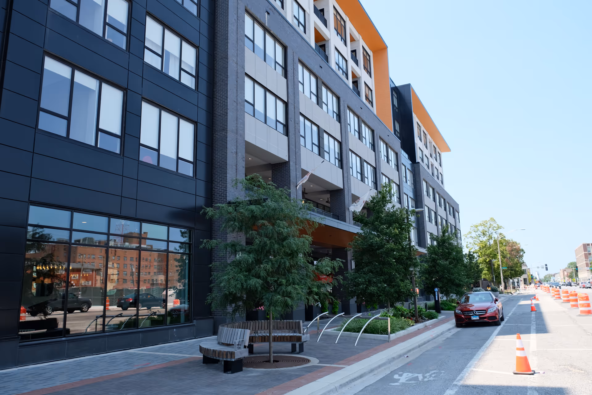 Exterior view of a modern multi-story building with large windows, trees, and benches along the sidewalk. A red car is parked on the street next to orange traffic cones.