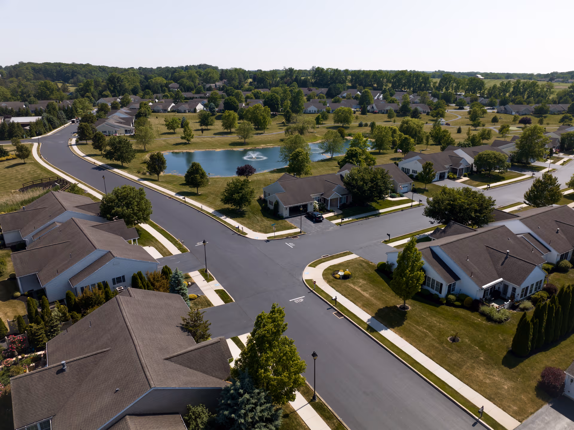 Aerial view of a residential neighborhood with single-story houses, well-maintained lawns, trees, and a small pond with a fountain in the center. Roads and sidewalks are visible, and the area is surrounded by greenery and open fields.