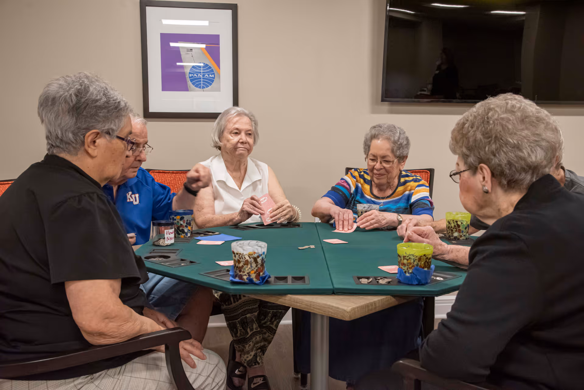 A group of elderly people sitting around a green card table playing cards in a room with beige walls. There is a framed picture on the wall and a large flat-screen TV mounted above it. Each person has a colorful cup in front of them.