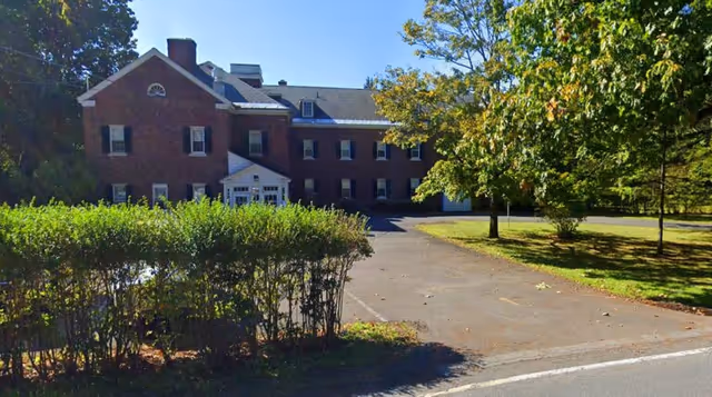 A large two-story brick building with white trim, surrounded by trees and greenery. There is a paved driveway leading up to the building, and a hedge in the foreground. The sky is clear and blue.