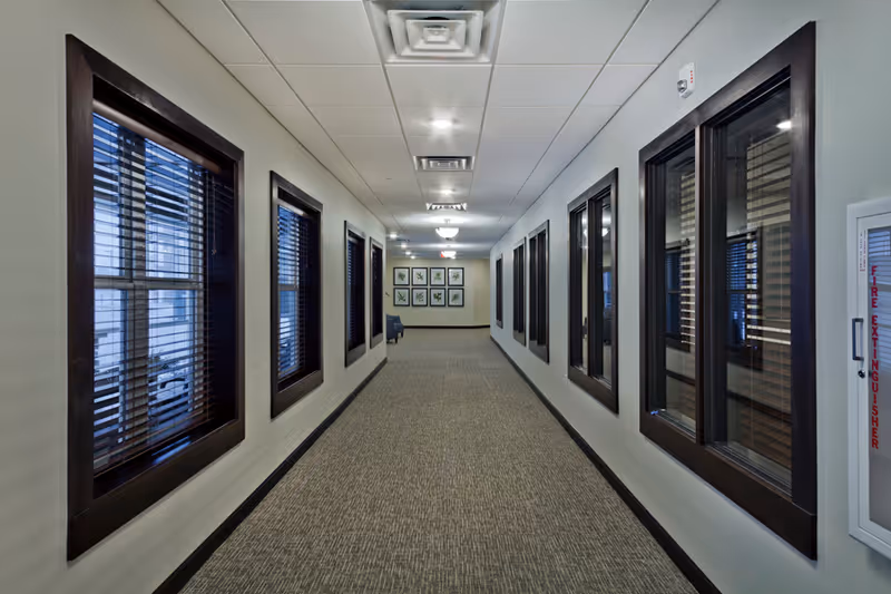 A long, carpeted hallway with multiple windows on both sides featuring dark wooden frames and blinds. The ceiling has recessed lighting and ventilation panels. At the end of the hallway, there are framed botanical prints on the wall and a blue chair. A fire extinguisher cabinet is mounted on the right wall.