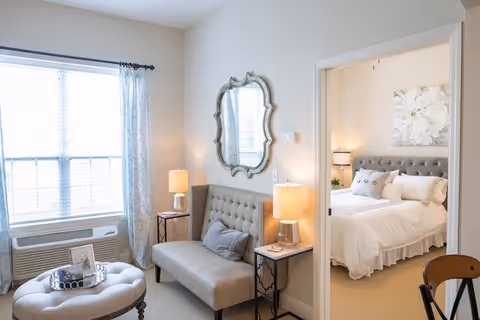A bright and cozy living area with a tufted loveseat, two side tables with lamps, a decorative mirror on the wall, and a large window with curtains. Through an open doorway, a bedroom with a neatly made bed, pillows, a nightstand with a lamp, and a floral wall art is visible.
