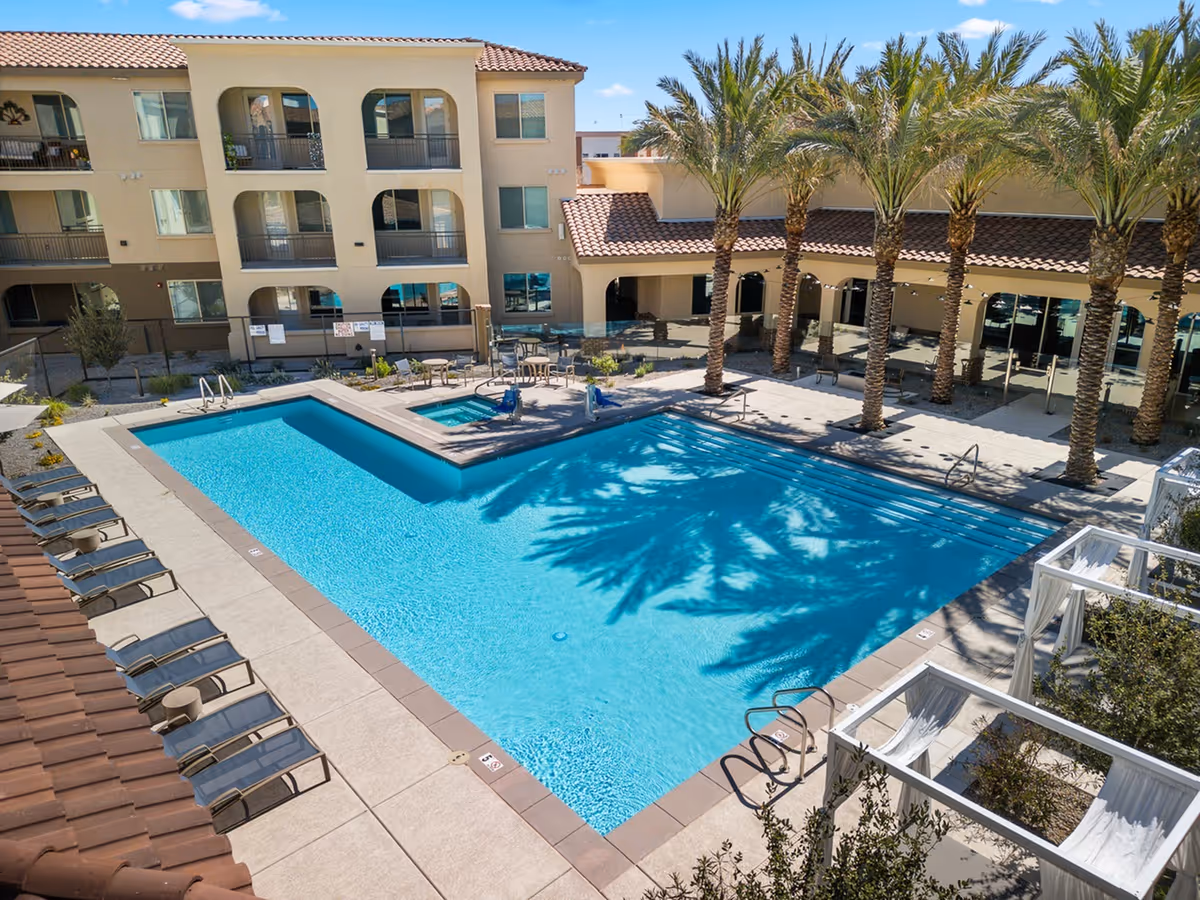 Outdoor swimming pool area at Album Marana facility with clear blue water, surrounded by lounge chairs, palm trees, and a building with balconies and arched windows in the background under a clear blue sky.