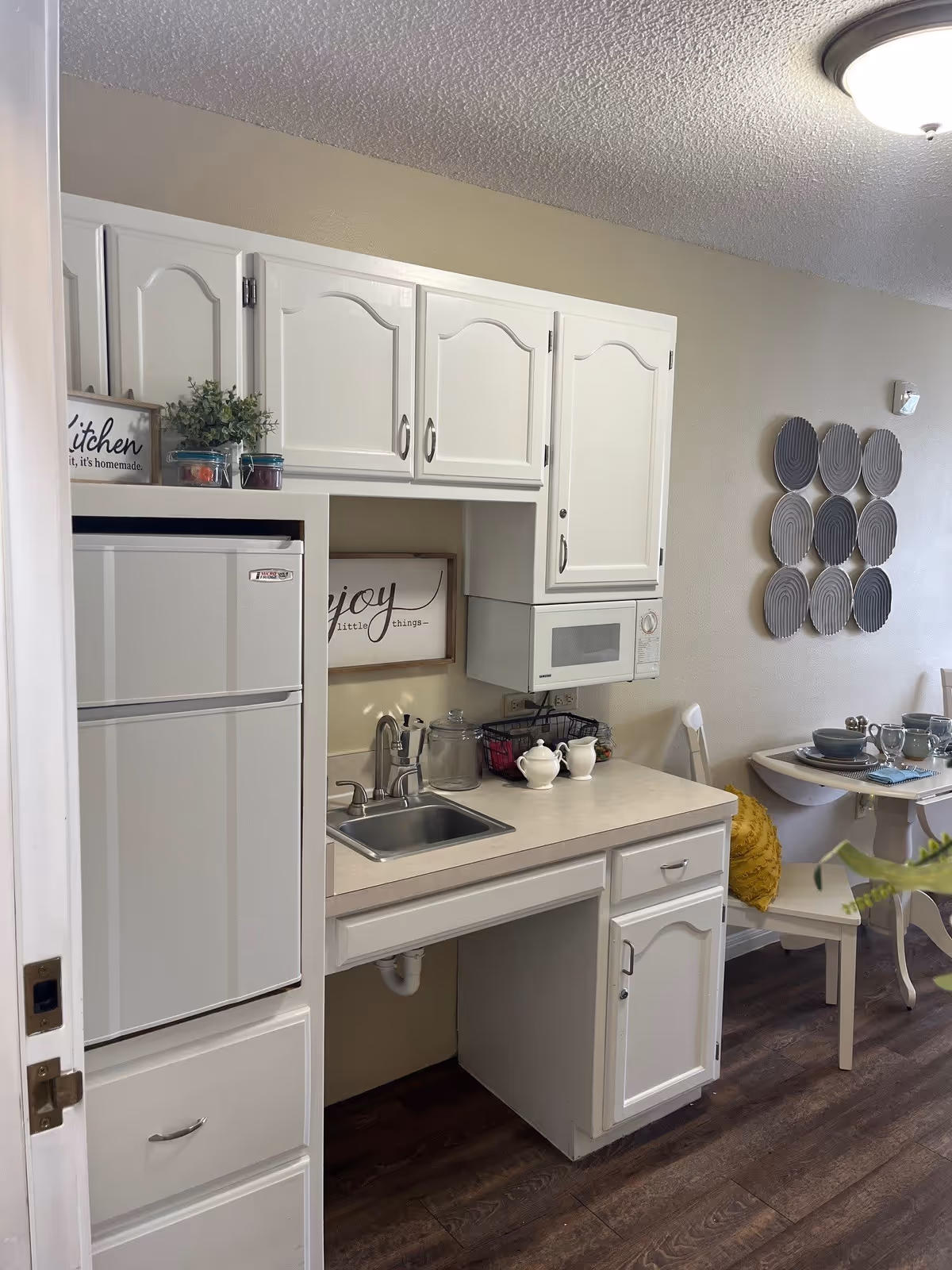 A small kitchen area with white cabinets, a countertop with a sink, a microwave mounted above the counter, and a white refrigerator. There are decorative signs on the wall and countertop, a small plant, and some kitchenware. To the right, a dining table is set with dishes and glasses, accompanied by white chairs, one with a yellow cushion. The floor is dark wood, and the walls are beige with a circular wall decoration.