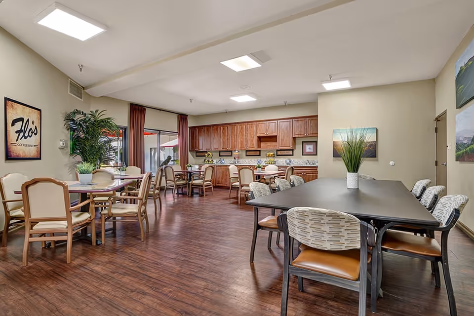 A spacious dining area with wooden flooring, multiple tables and chairs, and a kitchen counter with wooden cabinets in the background. The room features a sign that reads 'Flo's Coffee Bar', potted plants, and large windows with red curtains letting in natural light.