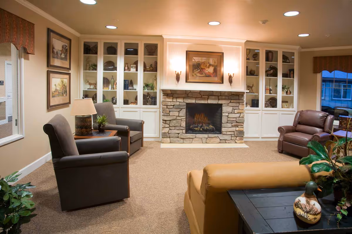 A cozy living room with a stone fireplace in the center, flanked by built-in white shelves displaying decorative items. The room has beige walls and carpet, with several comfortable chairs and a sofa arranged around the fireplace. There are framed pictures on the wall and a table with a lamp and plant.