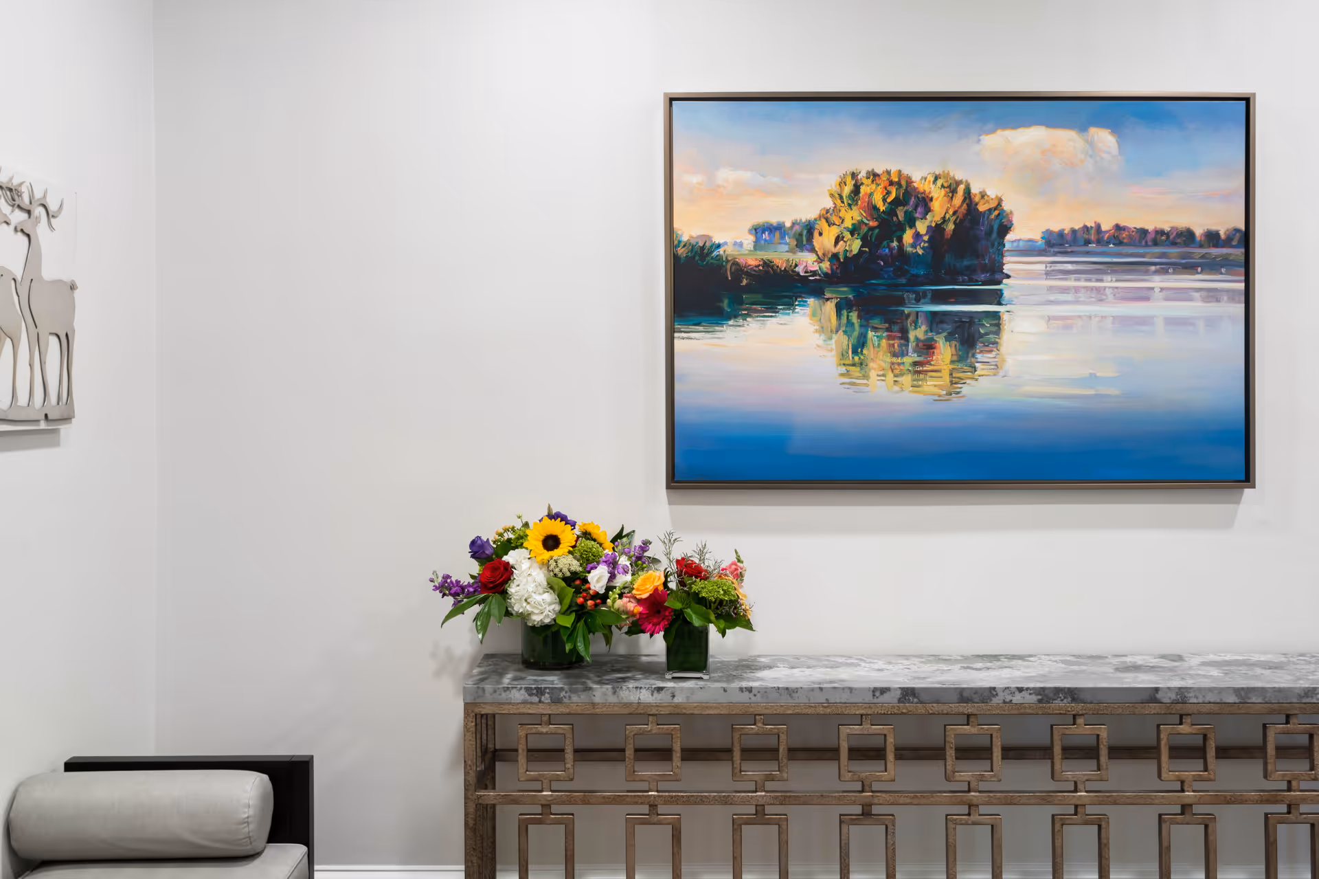 A modern interior corner featuring a decorative console table with a geometric metal base and a marble top. On the table are two vases with colorful flower arrangements. Above the table hangs a large framed painting of a serene water scene with trees reflected in the water. To the left, part of a light gray cushioned chair and a wall decoration with deer silhouettes are visible.