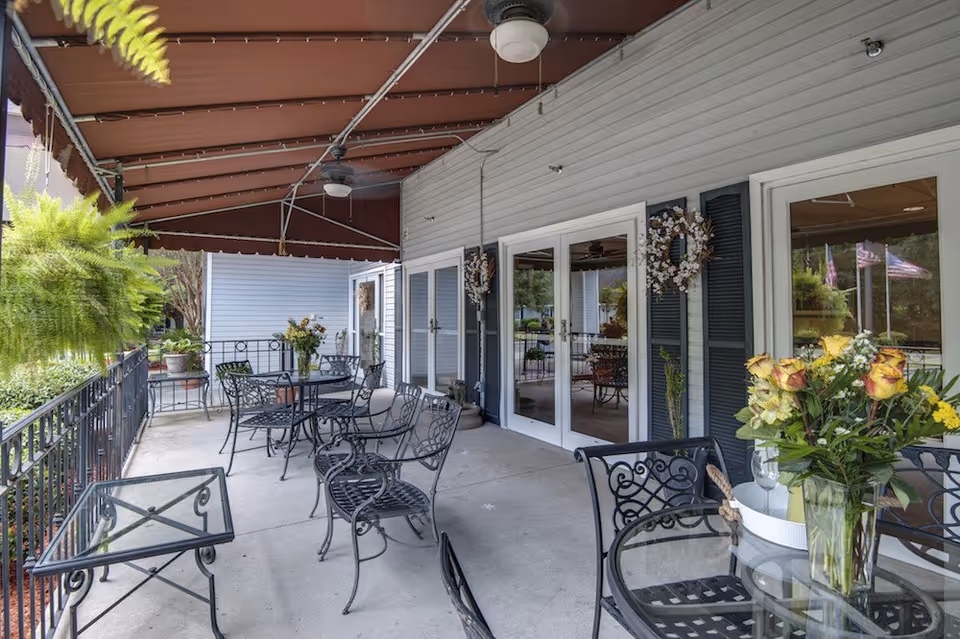 Covered outdoor patio with metal tables and chairs, potted plants and floral arrangements in front of glass doors.