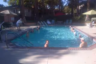 Outdoor swimming pool with several people in the water and a person standing near the poolside. Surrounding the pool are lounge chairs, umbrellas, and trees in the background.