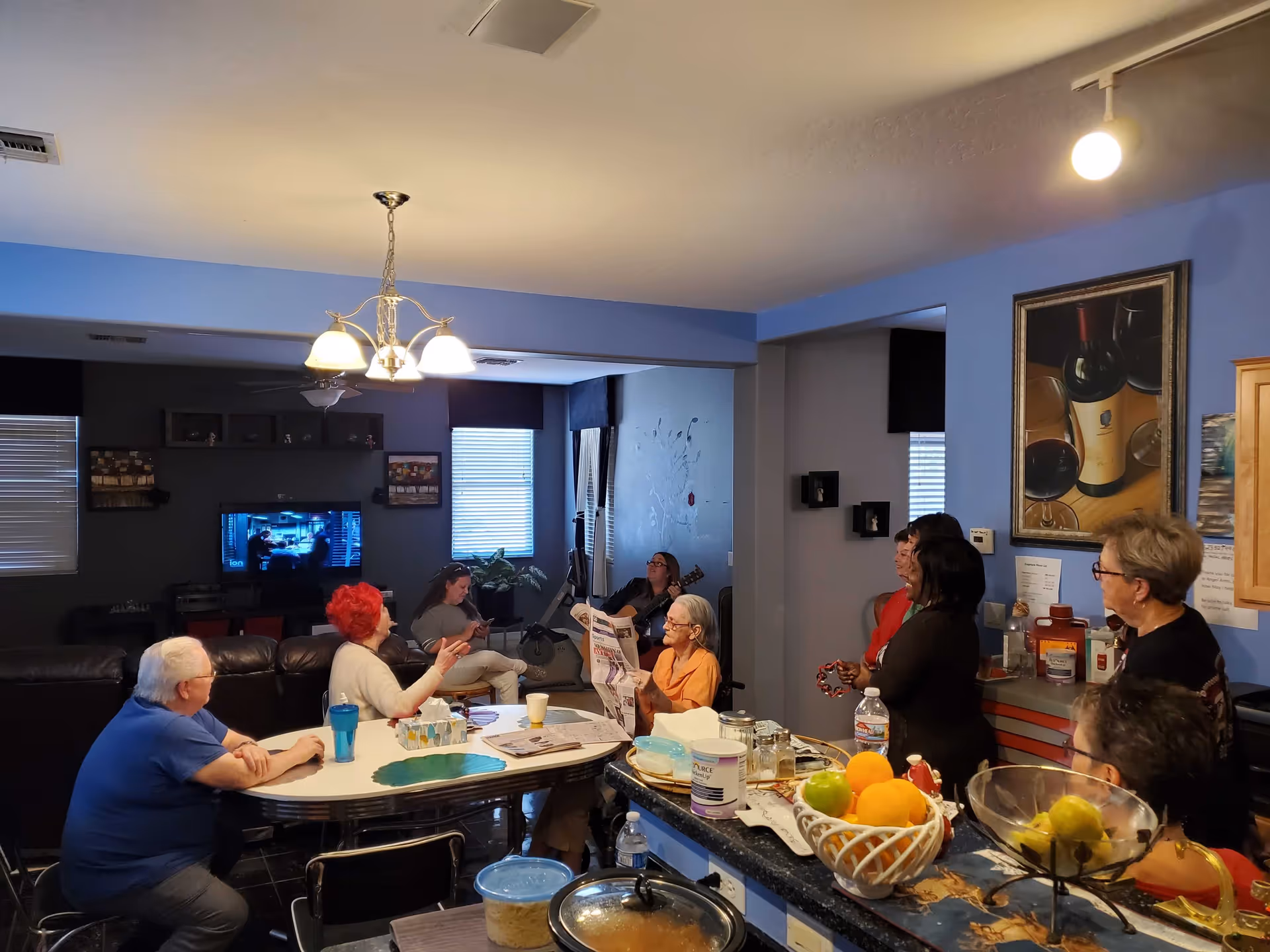 A group of elderly people and caregivers gathered in a cozy living room and kitchen area. Some are seated around a table with cups and snacks, while others stand near the kitchen counter with fruit bowls and kitchen items. A television is on in the background, and the room is warmly lit with a chandelier and ceiling light. The walls are decorated with paintings and shelves.