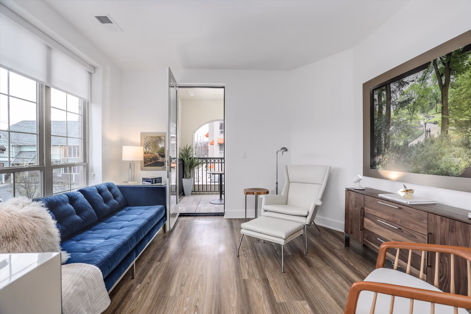 A modern living room with a blue velvet sofa on the left, a white armchair with an ottoman on the right, and a wooden sideboard with a large framed picture of a forest scene above it. The room has wooden flooring, white walls, and a door leading to a balcony with outdoor seating. A floor lamp and a small wooden side table are also visible.