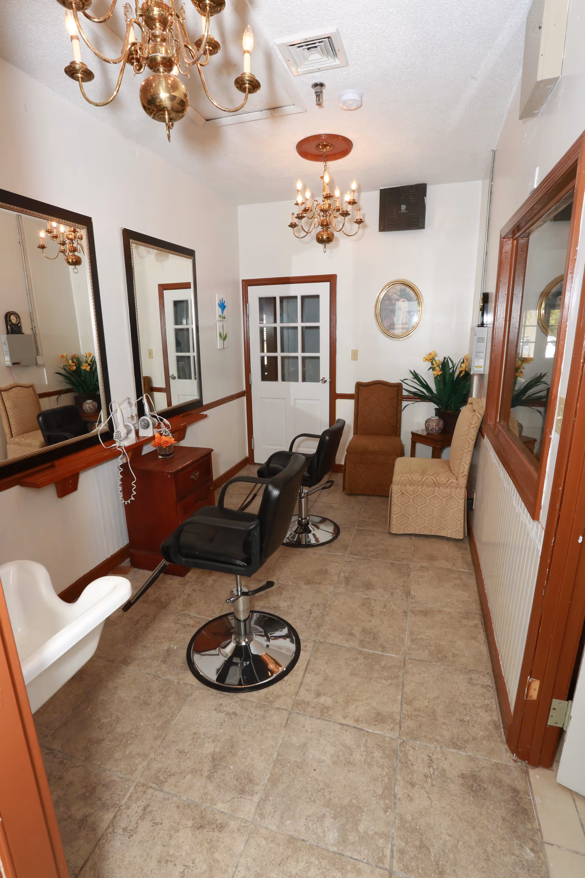 Interior salon area with two black styling chairs facing mirrors, chandeliers overhead, and seating at the back of a narrow room.