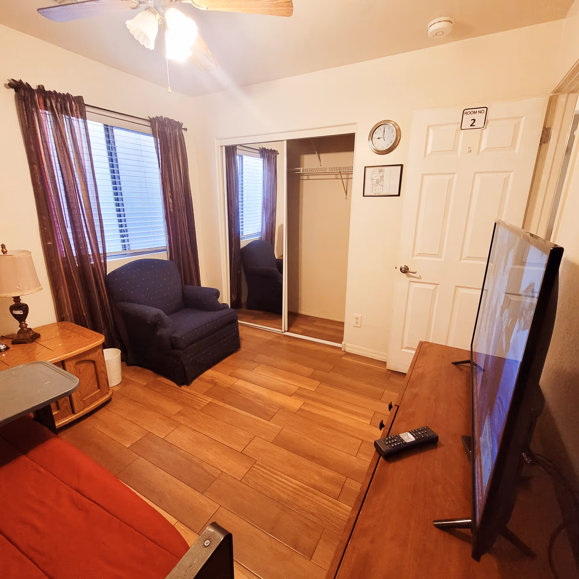 A small living room with wooden flooring, a blue armchair near a window with purple curtains, a wooden side table with a lamp, a wall clock, a door labeled 'Room No. 2', a TV on a wooden stand with a remote control, and a closet with sliding mirrored doors.