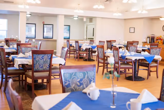 A bright and spacious dining room with multiple wooden tables and chairs. Each table is covered with white and blue tablecloths and set with white napkins and cups. The room has wooden flooring, several windows, and ceiling lights providing ample illumination.