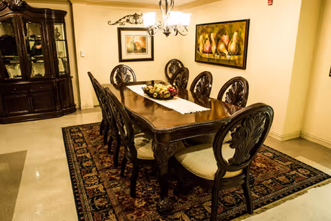 A formal dining room with a long wooden dining table surrounded by eight ornate wooden chairs with cushioned seats. The table has a white runner and a centerpiece bowl with decorative fruit. The room features a patterned area rug under the table, a chandelier overhead, a large wooden china cabinet against the wall, and framed artwork on the walls.