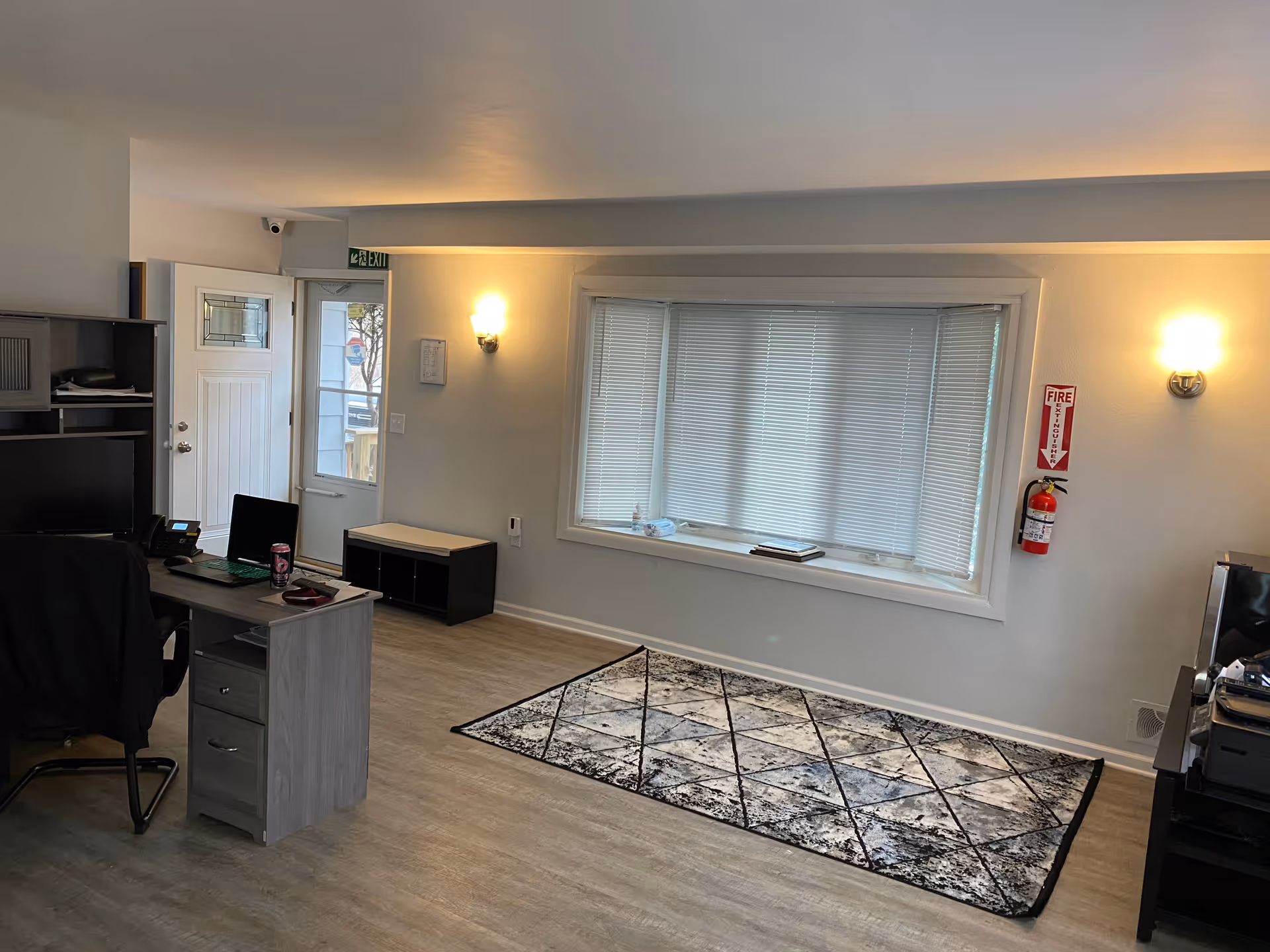 Interior view of a room with light-colored walls and flooring, featuring a large window with closed blinds, a patterned area rug on the floor, a desk with a computer and office chair, a small bench near the door, and wall-mounted lights on either side of the window. A fire extinguisher is mounted on the wall to the right of the window.