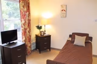 A small bedroom with a single bed covered in a brown bedspread and a beige pillow. Next to the bed is a dark wooden nightstand with a lamp, a small plant, and a framed photo. Another dark wooden dresser with a small flat-screen TV on top is positioned near a window with red and gold patterned curtains. The walls are light-colored, and the room has a calm, simple atmosphere.