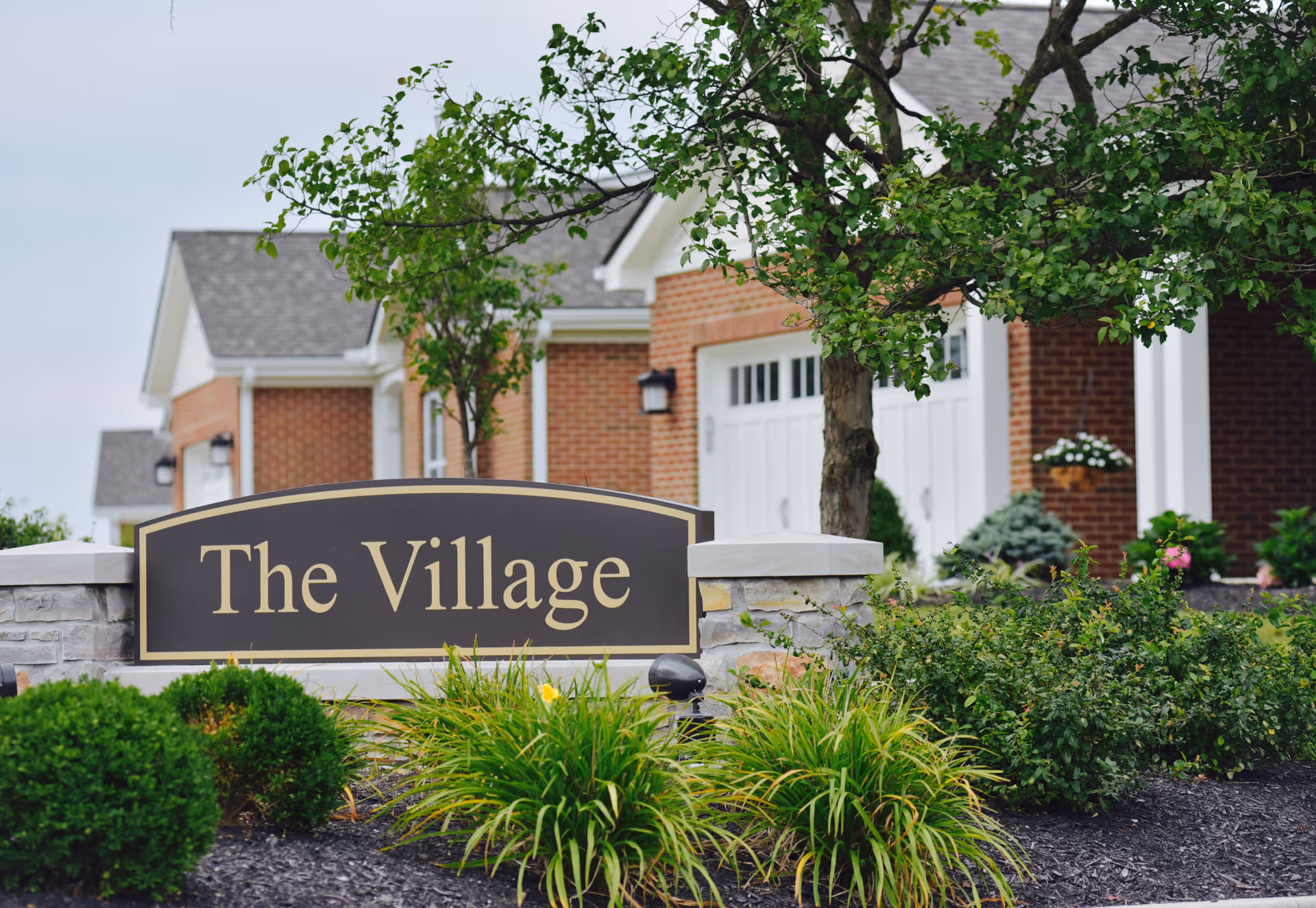 A landscaped entrance area with a sign that reads 'The Village' in front of brick buildings and greenery, including bushes and a tree.