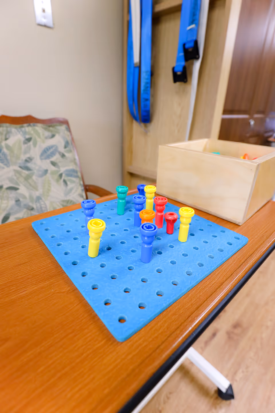 A close-up view of a wooden table with a blue pegboard and colorful pegs arranged on it. Behind the table, there is a wooden box containing more pegs, a chair with a floral patterned cushion, and some blue straps hanging on a wooden panel in the background.