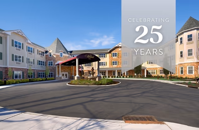 Exterior view of The Bristal Assisted Living at Lake Grove building with a circular driveway and a covered entrance. The building has multiple windows, a mix of brick and siding exterior, and a clear blue sky above. A translucent banner on the right side reads 'Celebrating 25 Years'.