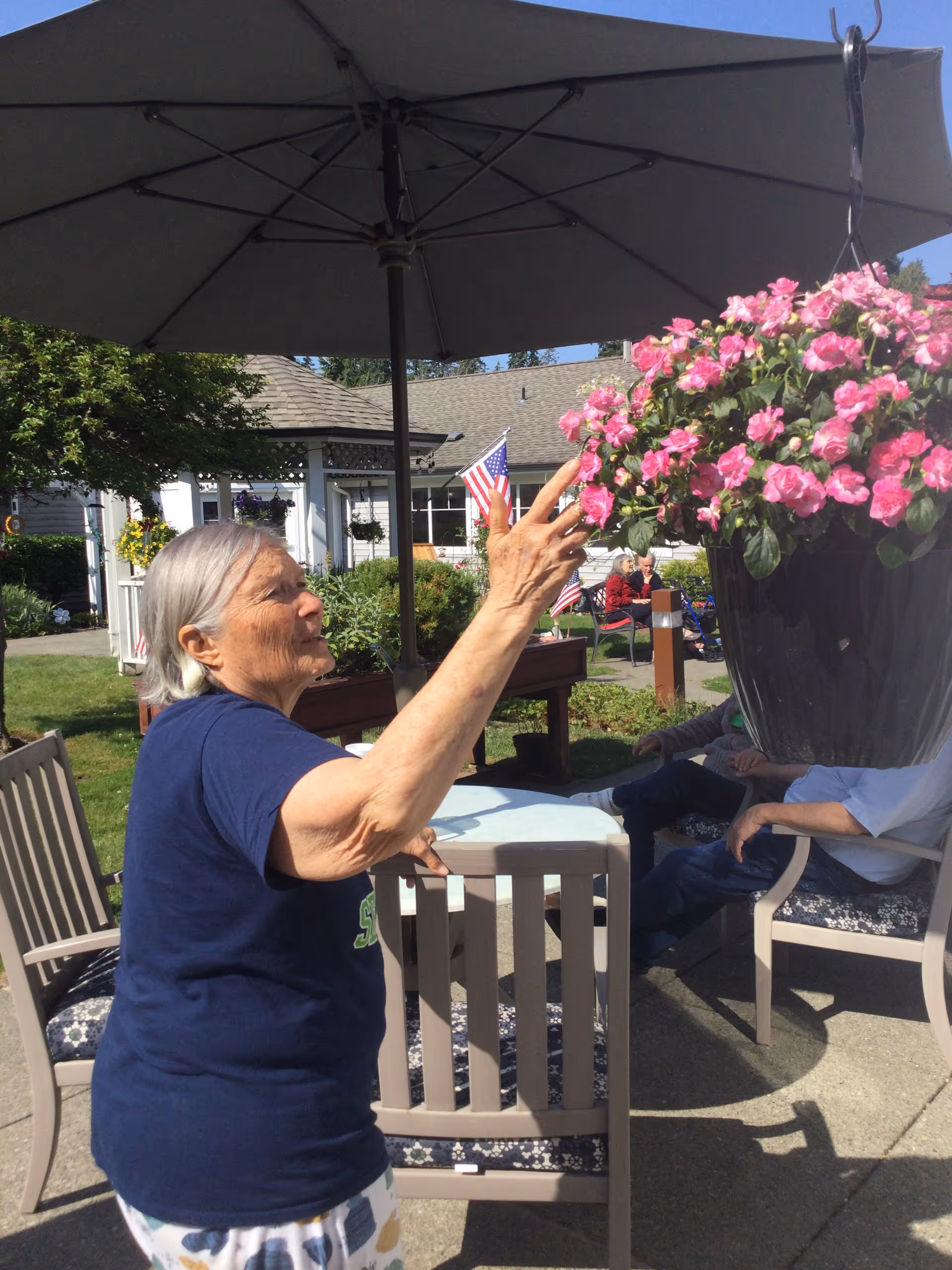 An elderly woman in a courtyard reaches to touch a hanging pot of pink flowers under a patio umbrella.