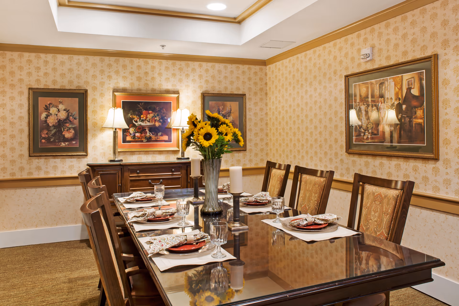 A formal dining room with a long glass-top wooden table set for six people. Each place setting has a red plate, a patterned napkin, and a glass. A vase with sunflowers and two candle holders with white candles are centered on the table. The walls are decorated with floral wallpaper and framed paintings, and there are two table lamps on a wooden sideboard against the wall.