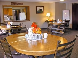 A dining area with round wooden tables and black metal chairs. Each table has a centerpiece with autumn-themed decorations and condiments. In the background, there is a kitchen pass-through window, a lamp on a side table, framed wall art, and a whiteboard. The room has beige walls and carpeted flooring.